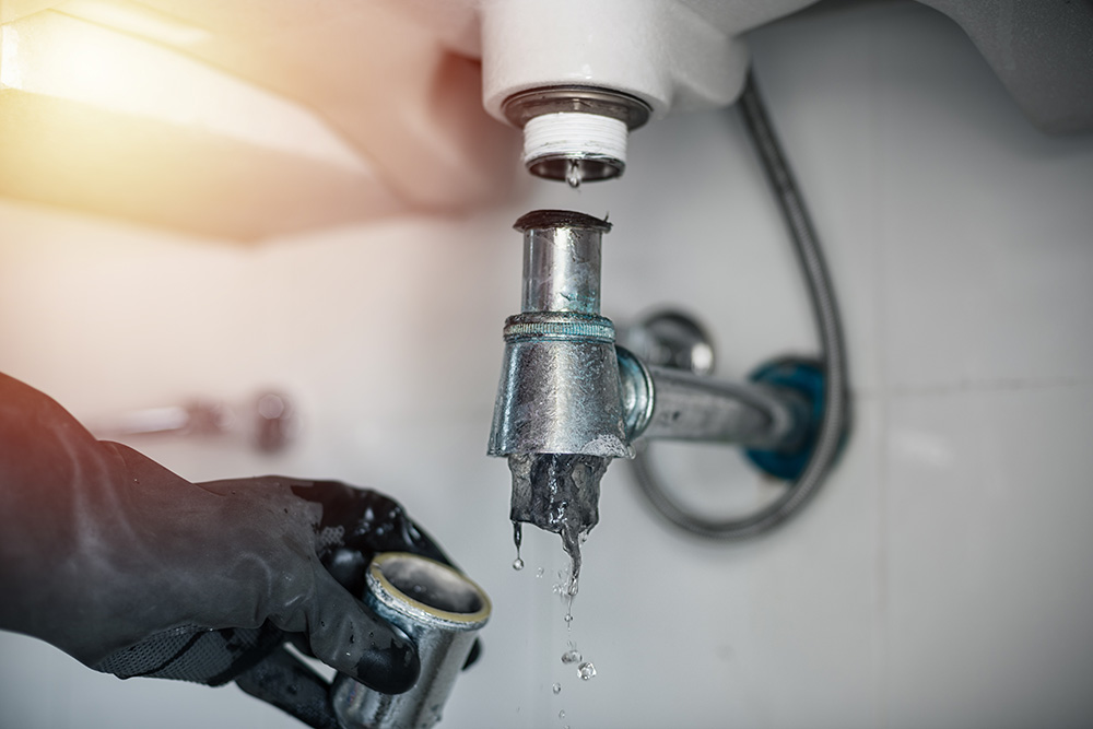 Omaha Home, Kitchen, Bathroom and Basement Remodeling - A person wearing a black glove adjusts a leaking silver pipe under a sink, with water dripping from the connection point.