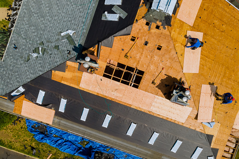 Omaha Home, Kitchen, Bathroom and Basement Remodeling - Aerial view of workers on a residential roof under construction, with sections covered in plywood and roofing materials, depicting a construction site in progress.