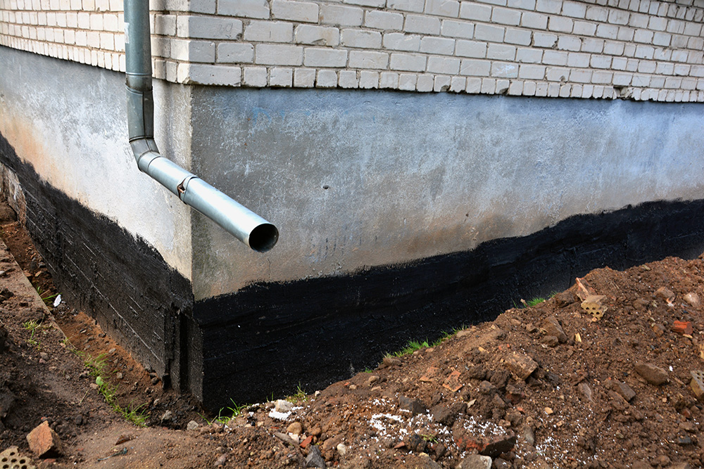Omaha Home, Kitchen, Bathroom and Basement Remodeling - A drainage pipe protruding from a building wall with waterproofing foundations, surrounded by exposed soil. the wall is partly painted in black and grey.
