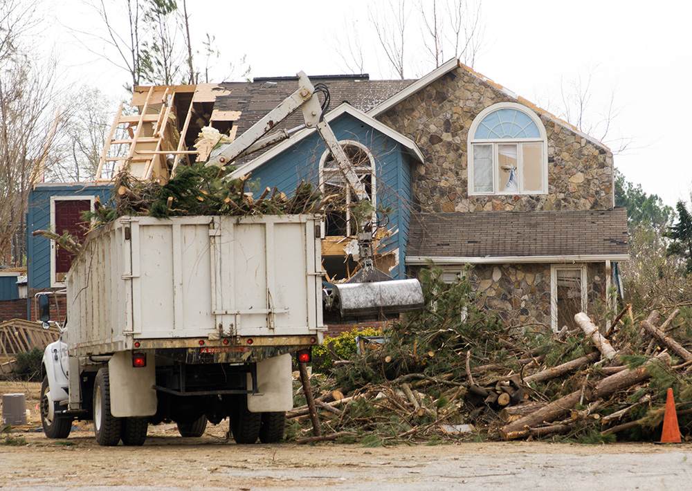 Omaha Home, Kitchen, Bathroom and Basement Remodeling - A dump truck loaded with debris parked in front of a heavily damaged house with a destroyed roof and scattered tree branches around, indicating recent storm damage.