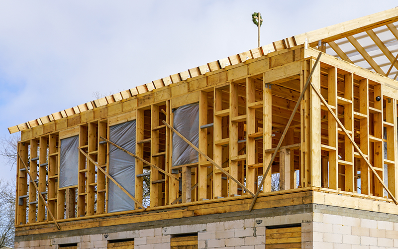 Omaha Home Additions, Kitchen, Bathroom and Basement Remodeling - An under-construction building with wooden framing, partial roofing, and a brick base. The structure shows exposed wooden beams and plastic-covered window openings. Clear sky is in the background.