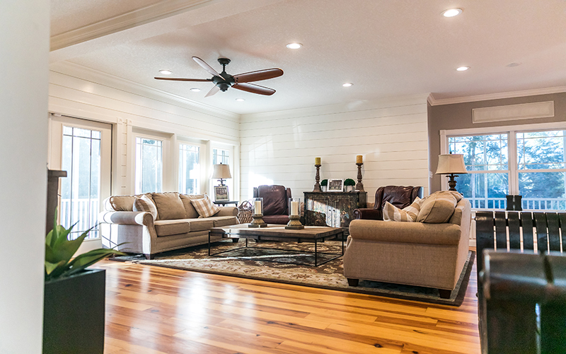 Omaha Home additions, Kitchen, Bathroom and Basement Remodeling - A cozy living room with large windows, off-white walls, a ceiling fan, and hardwood floors. It features two beige sofas, two brown armchairs, a coffee table, and a fireplace with wooden detailing, candles, and decorative pieces. A green plant is in the foreground.