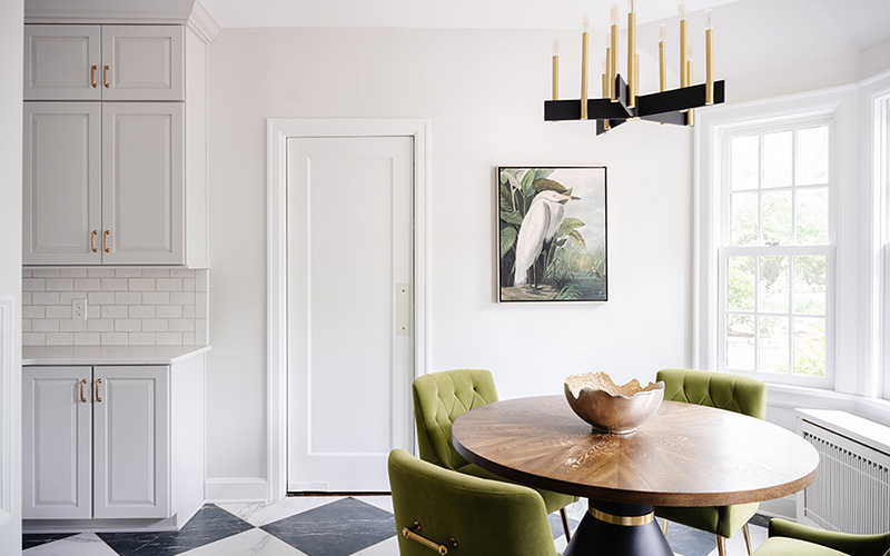 Omaha Home, Kitchen, Bathroom and Basement Remodeling - A bright and elegant dining area with a wooden round table, green upholstered chairs, and a chic chandelier, featuring white cabinetry, subway tile backsplash, and a painting of birds on the wall.