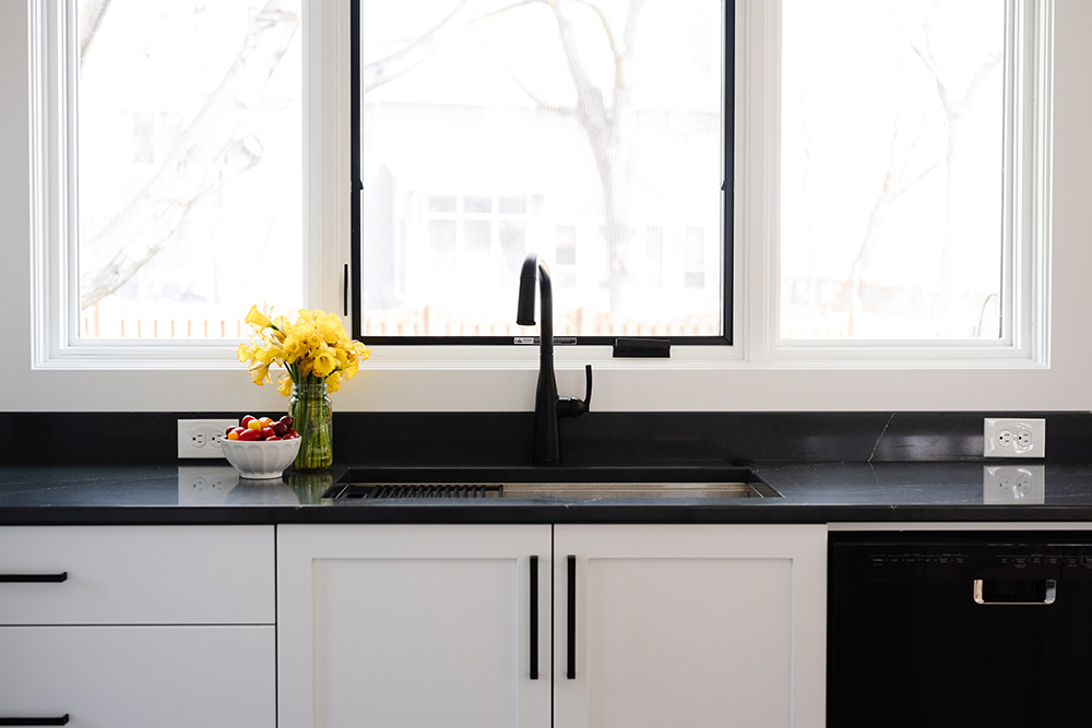 Omaha Home, Kitchen, Bathroom and Basement Remodeling - A modern kitchen renovation with black countertops and white cabinets, featuring a sink with a tall faucet. A bouquet of yellow flowers and a bowl of red and yellow fruits enhance the space, with a window view of bare trees.
