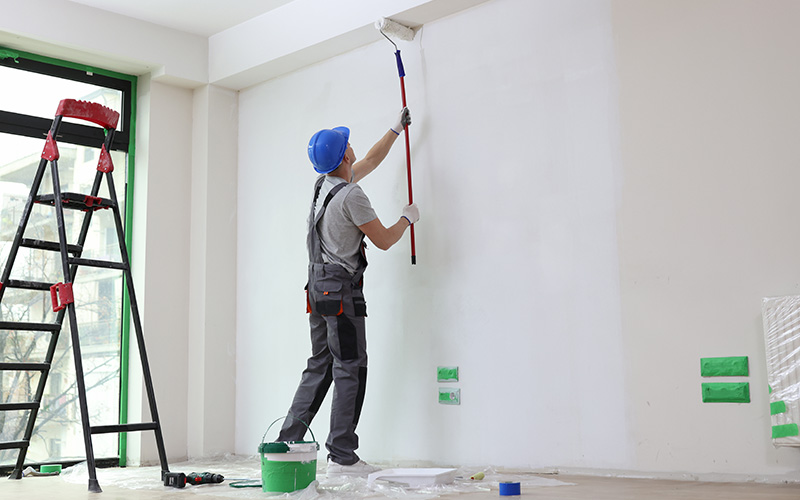 Omaha Home, Kitchen, Bathroom and Basement Remodeling - A painter in a blue hard hat and work overalls is using a roller to apply white paint to a wall, with painting equipment and a ladder nearby.